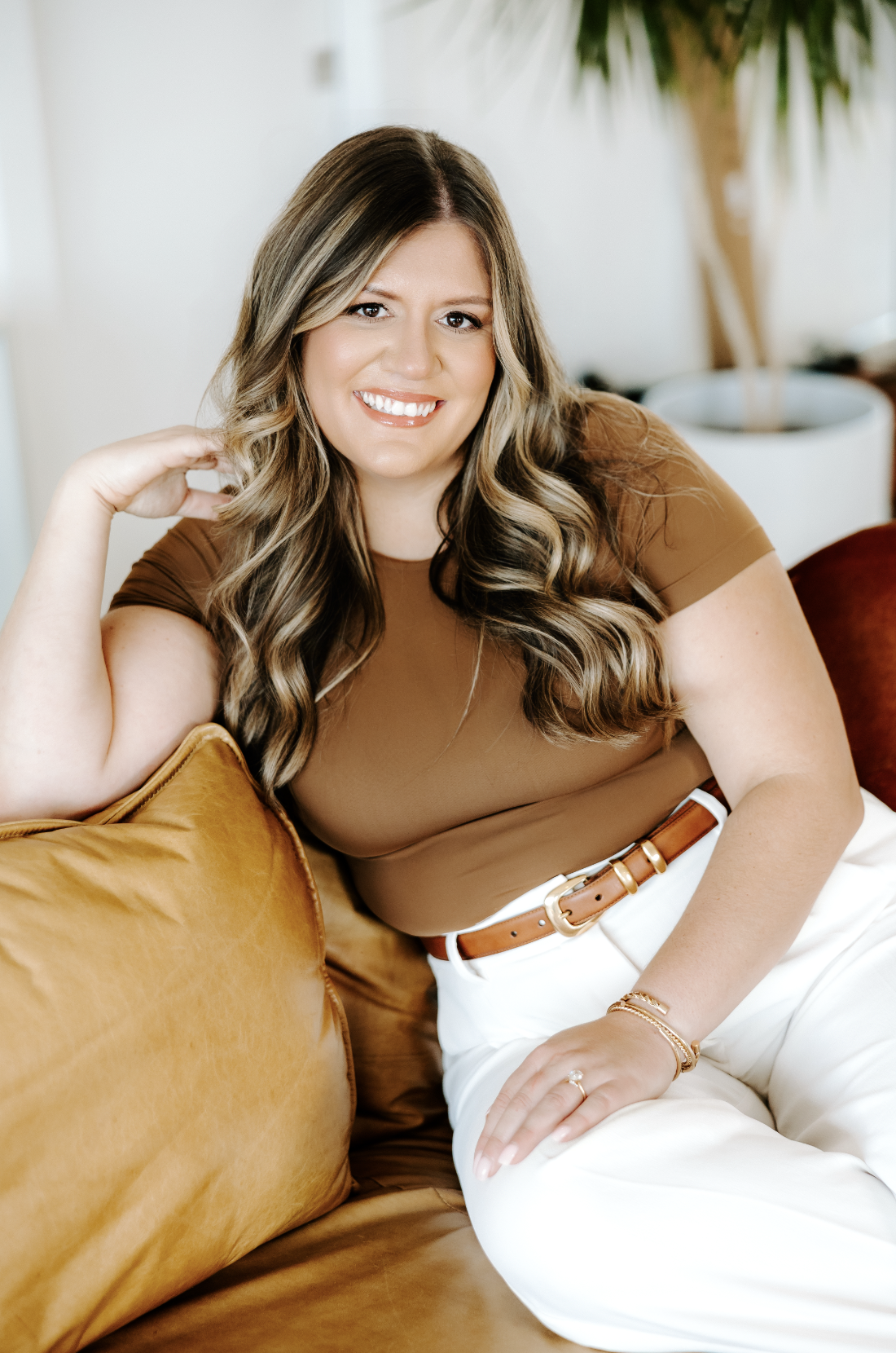 Heather Gorr, LPC-S, trauma-informed therapist and clinical supervisor based in Austin, Texas | Woman with dark curly hair smiling, wearing a brown shirt and white pants, sitting on a brown leather couch