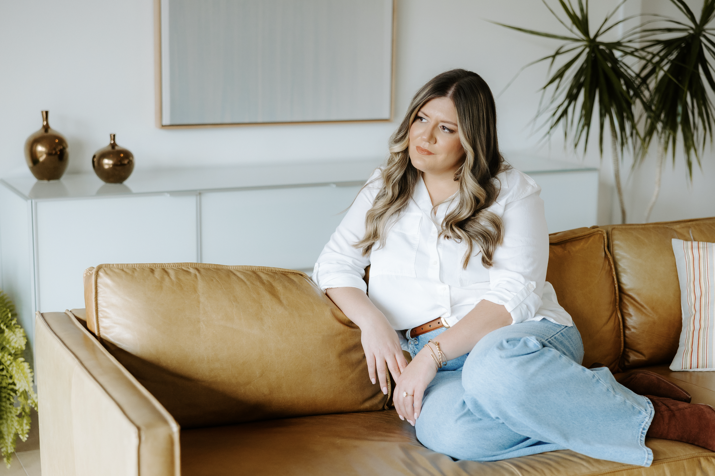 Heather Gorr, LPC-S, trauma-informed therapist and clinical supervisor based in Austin, Texas | Woman with dark curly hair, wearing a white shirt and blue jeans, sitting on a brown leather couch