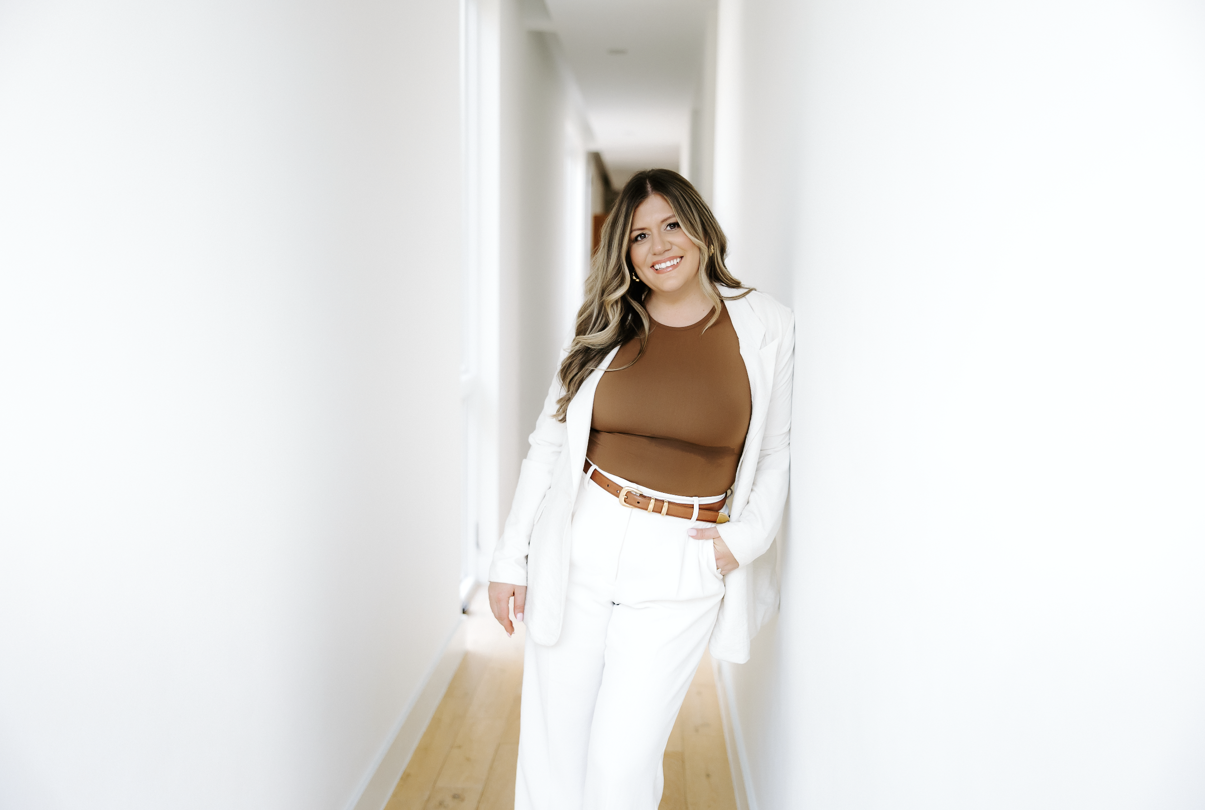 Woman in white pants and blazer standing in a bright hallway, smiling at the camera.