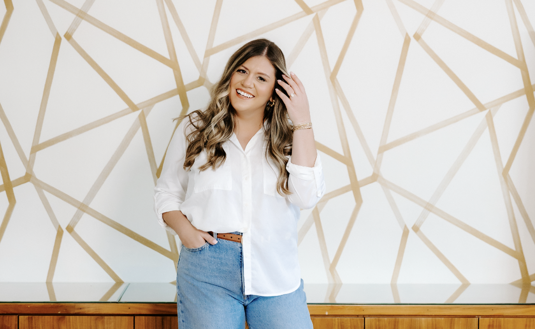 A woman with long wavy hair smiling, wearing a white shirt and blue jeans, standing in front of a cream wall with a gold geometric pattern.