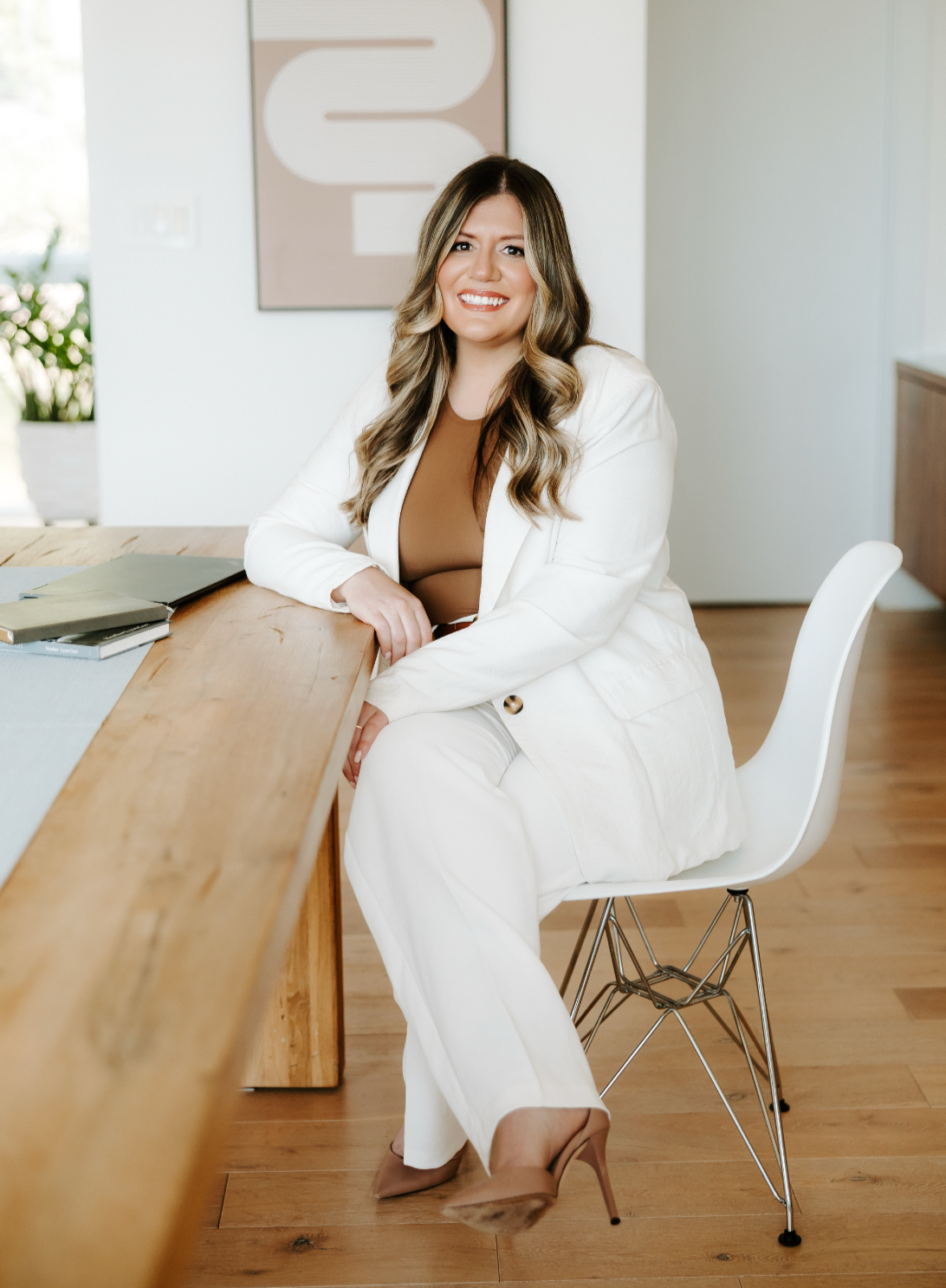 Heather Gorr, LPC-S, trauma-informed therapist and clinical supervisor based in Austin, TexasA woman with long wavy hair, wearing a white suit, smiling while sitting in a modern office room with a wooden floor, white walls, and a desk with notebooks.