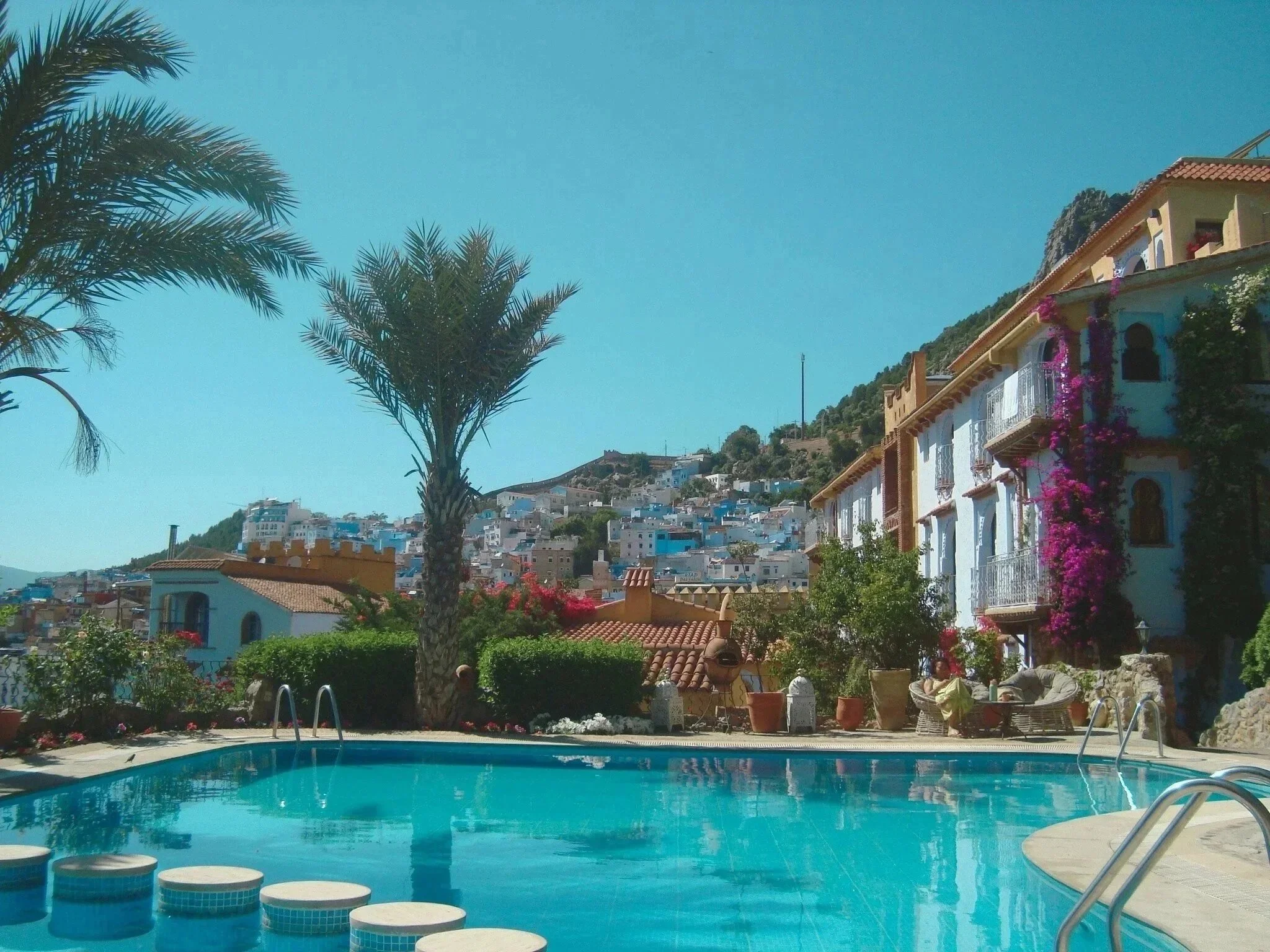 Swimming pool with palm trees and colorful buildings in a hillside town under clear blue sky.