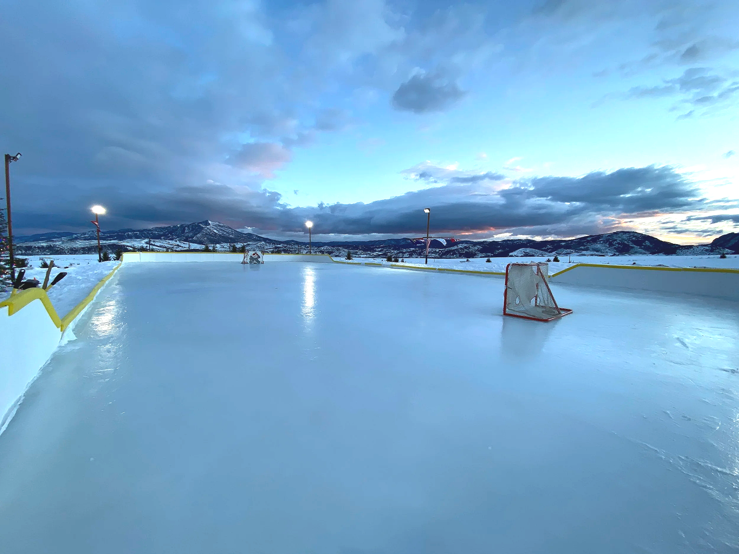 An outdoor ice hockey rink at dusk with mountains in the background, illuminated by lights, with two small hockey goals on the ice.