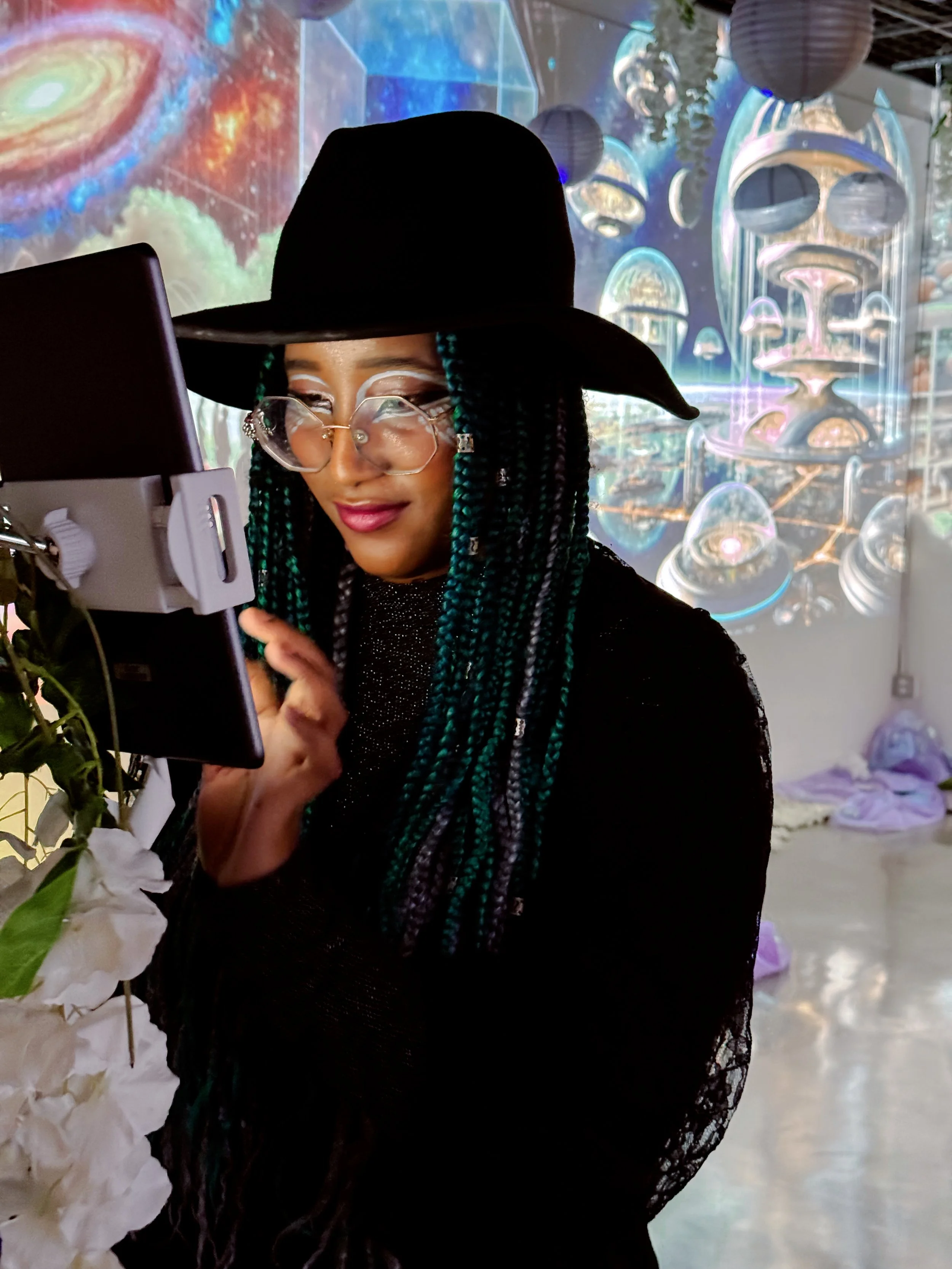 A woman with long braided hair, wearing glasses and a wide-brimmed black hat, looking at a tablet device in a colorful, space-themed art installation.