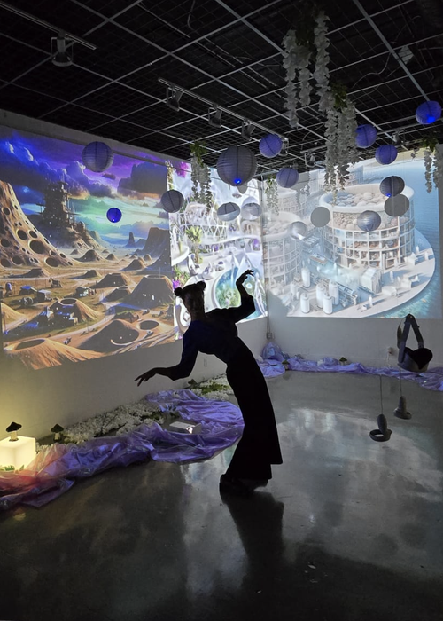 Person dancing in a futuristic, artistic space with space-themed projections on the walls, hanging paper lanterns, and decorative plants.
