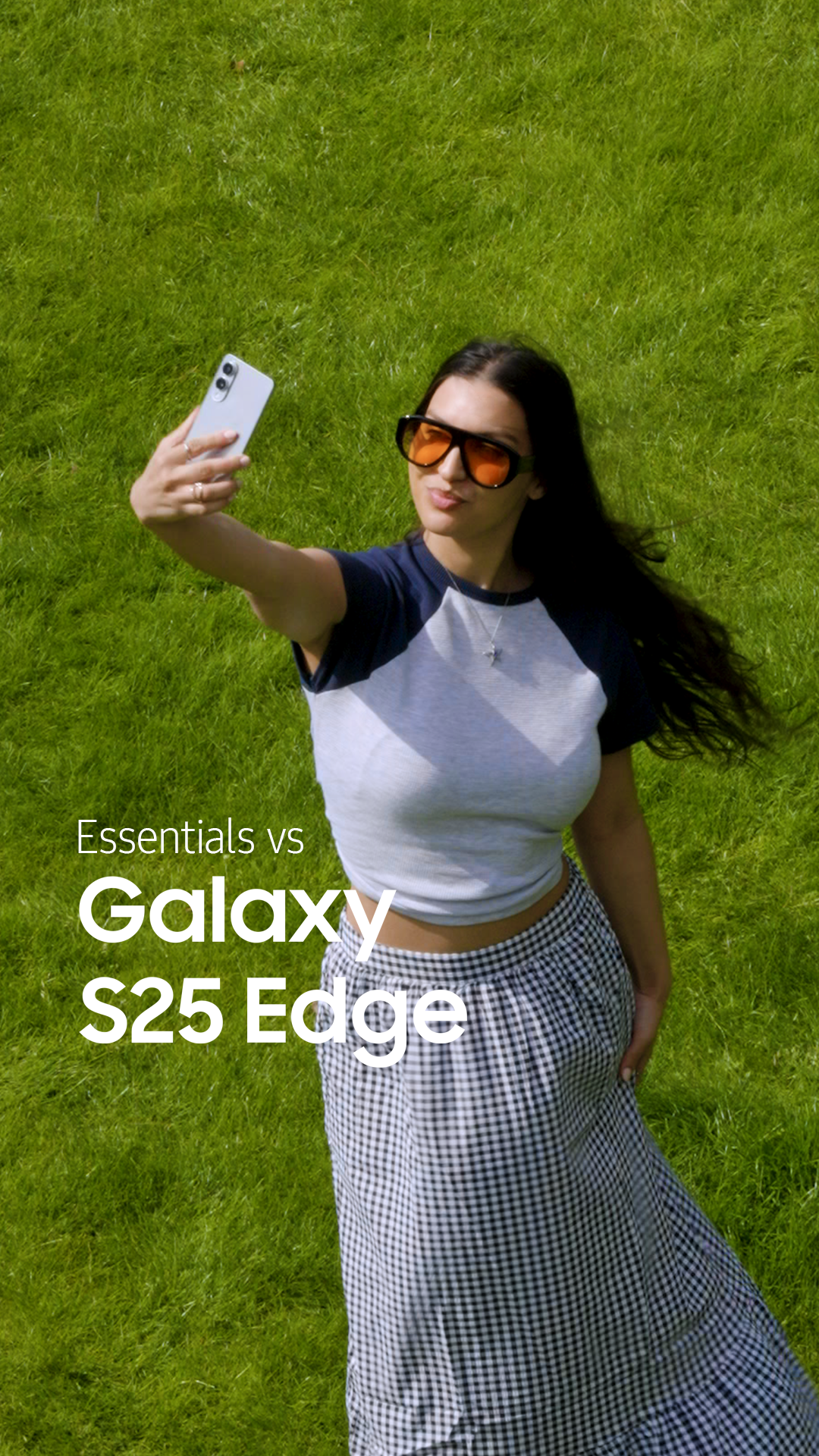 A woman taking a selfie outdoors on the grass, wearing sunglasses, a gray and navy t-shirt, and a checkered skirt. The text on the image reads 'Essentials vs Galaxy S25 Edge.'