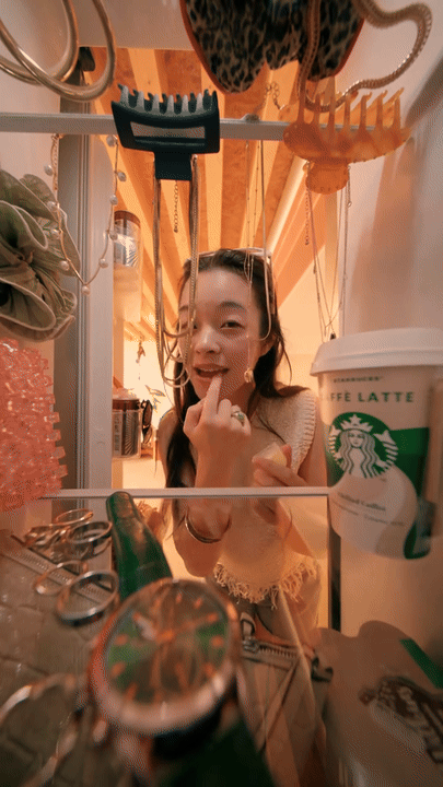 A young woman is looking into a fridge, smiling with a finger on her lips, surrounded by various kitchen items and a Starbucks iced coffee cup.