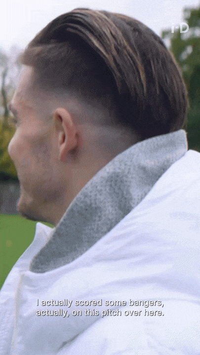 Close-up of a man with a fade haircut, wearing a white and gray jacket, outdoors on a soccer field.