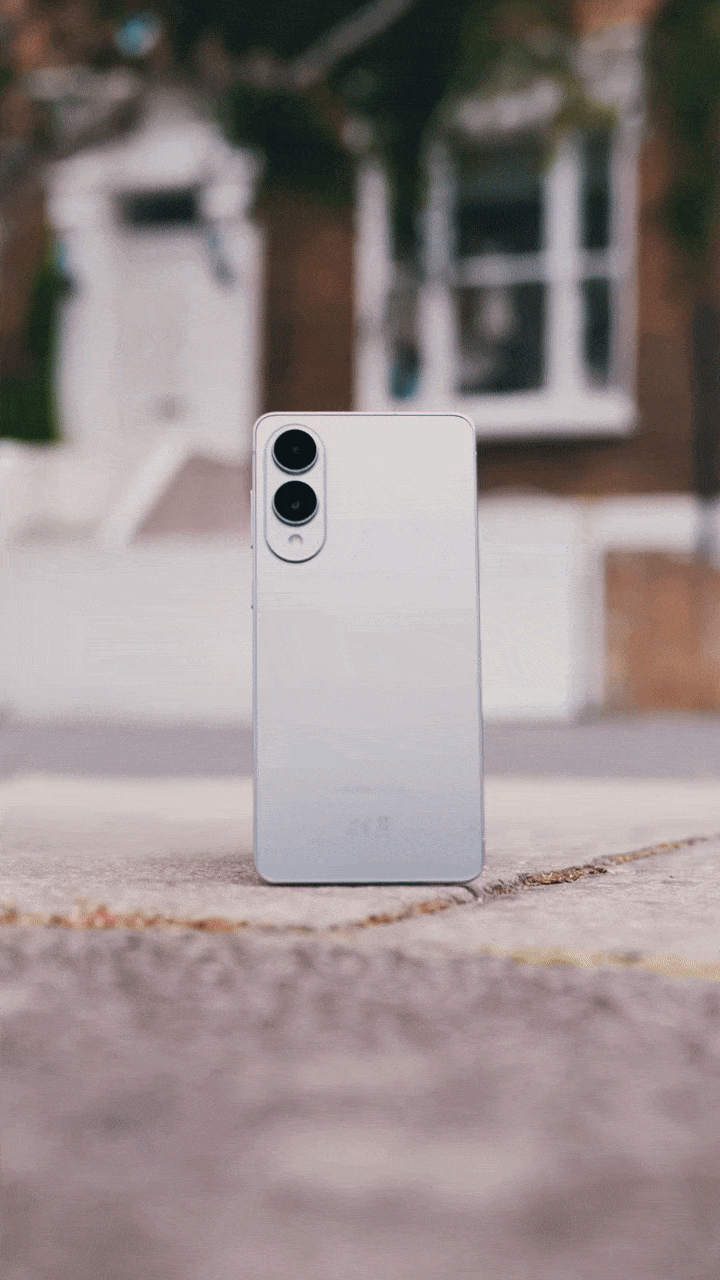 White smartphone with dual camera lenses and flash on a sidewalk in front of a house with a white window and brick wall.