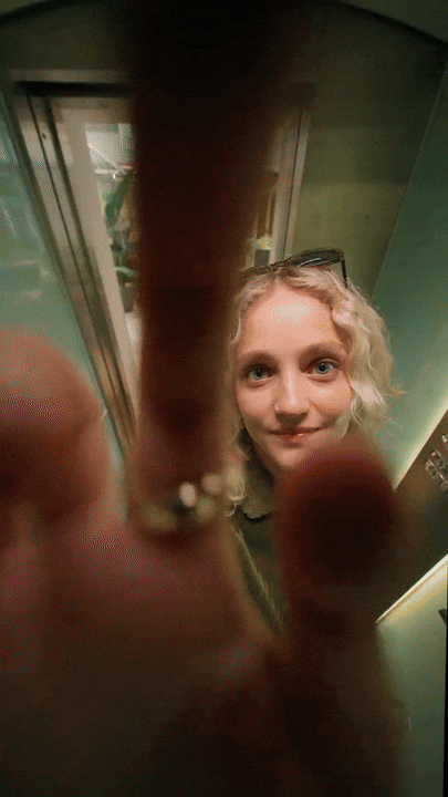 A girl with blonde curly hair taking a selfie in an elevator, holding the camera close to her face and partially covering it with her fingers.