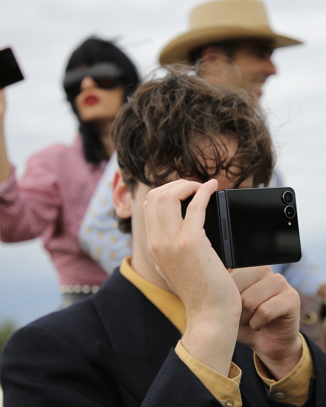 A group of people outdoors, with one person in the foreground taking a photo with a Samsung foldable smartphone. Others in the background are wearing sunglasses and unique outfits, including a woman with a black bob haircut, a wide-brimmed hat, and a woman with a red-and-white checkered shirt.