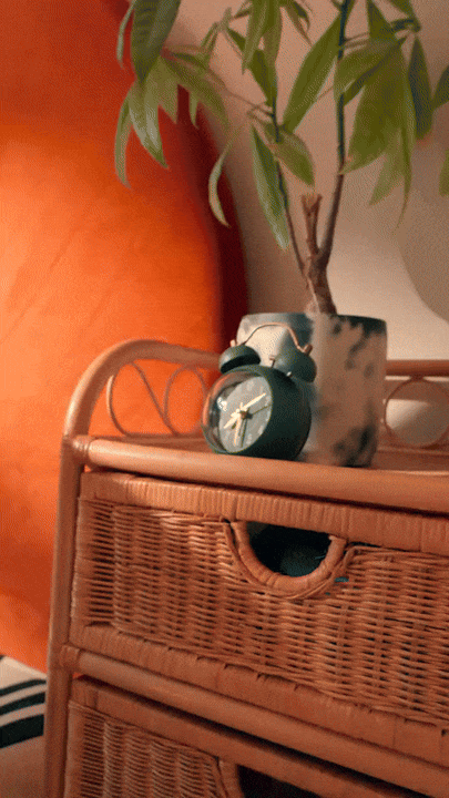 A small alarm clock sitting on a rattan side table next to a potted plant with green leaves against an orange wall.
