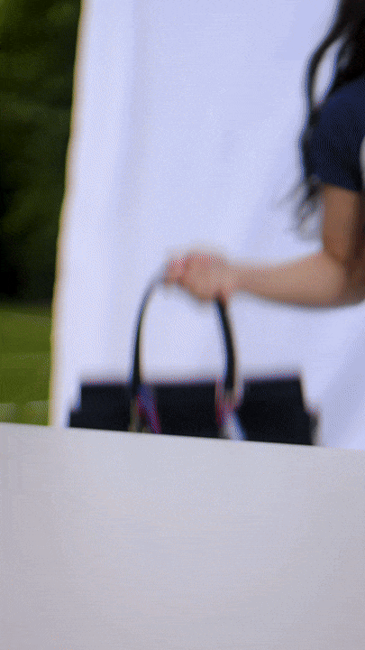 A woman holding a black handbag outdoors, with a white background and greenery in the distance.