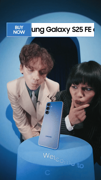 Two people looking at a Samsung Galaxy S25 FE smartphone displayed on a blue pedestal with a sign that says "Welcome to" and a banner in the background that says "BUY NOW Samsung Galaxy S25 FE".