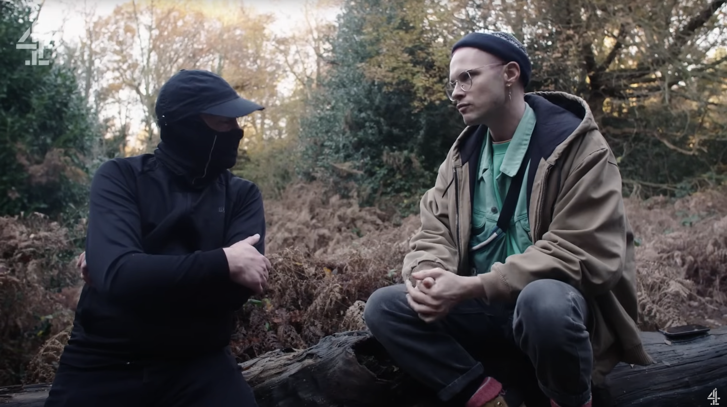 Two men sitting outdoors on a log in a wooded area with autumn foliage, engaged in a conversation.