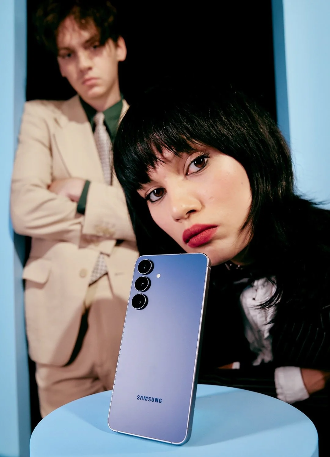 A woman with dark hair and red lipstick taking a selfie with a Samsung smartphone, while a man with light brown or dark blonde hair, wearing a beige suit and tie, stands in the background with his arms crossed in a dark blue booth.