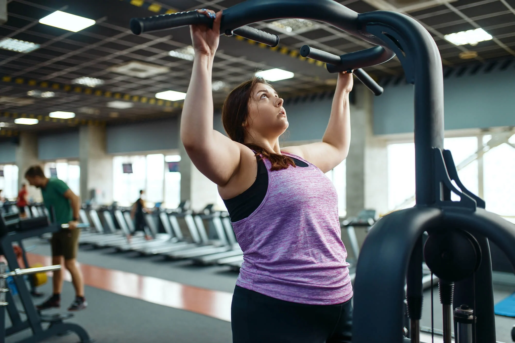 A woman lifting a pull-up bar in a gym with treadmills and other gym equipment in the background.