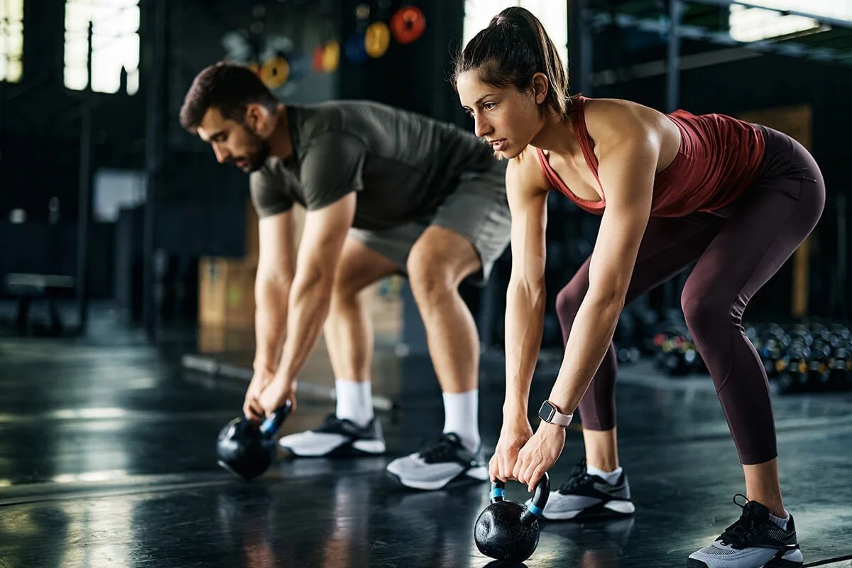 A man and a woman working out in a gym with kettlebells, both bent over and lifting kettlebells.