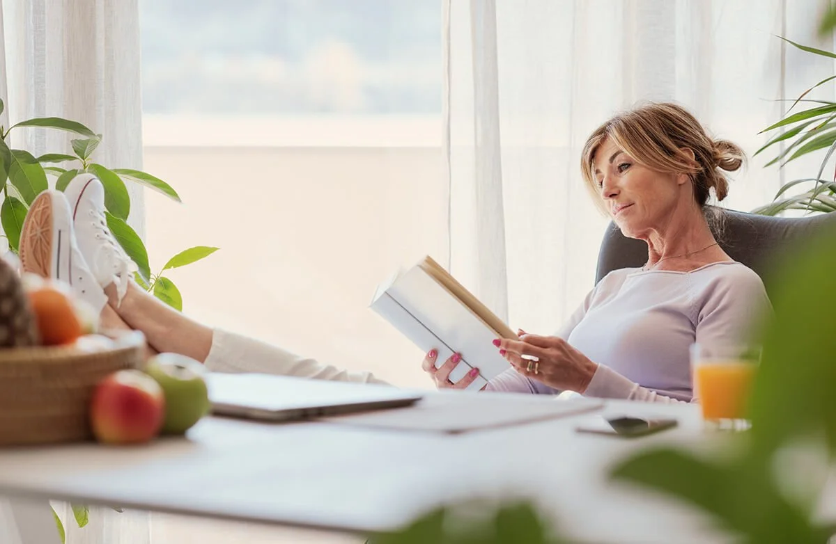 A woman with short hair relaxing in a bright room, sitting in a chair with her legs up on a table, reading a book, with natural light coming through a window behind her.