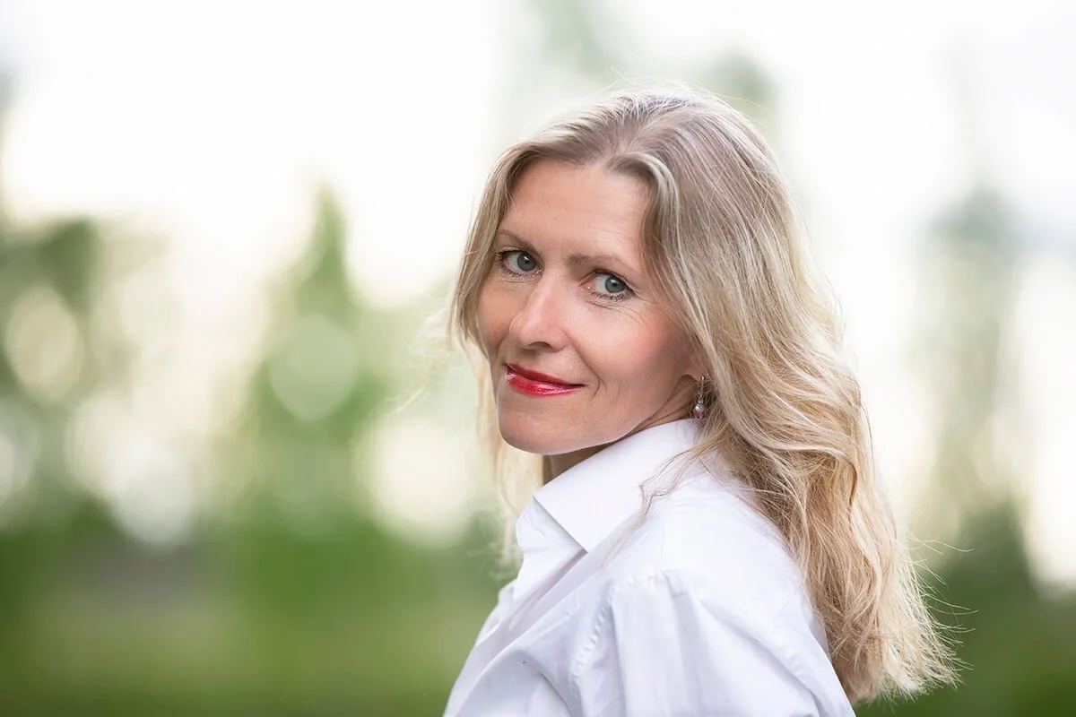 A woman with blonde hair, wearing a white shirt, smiling at the camera with a blurred outdoor background.