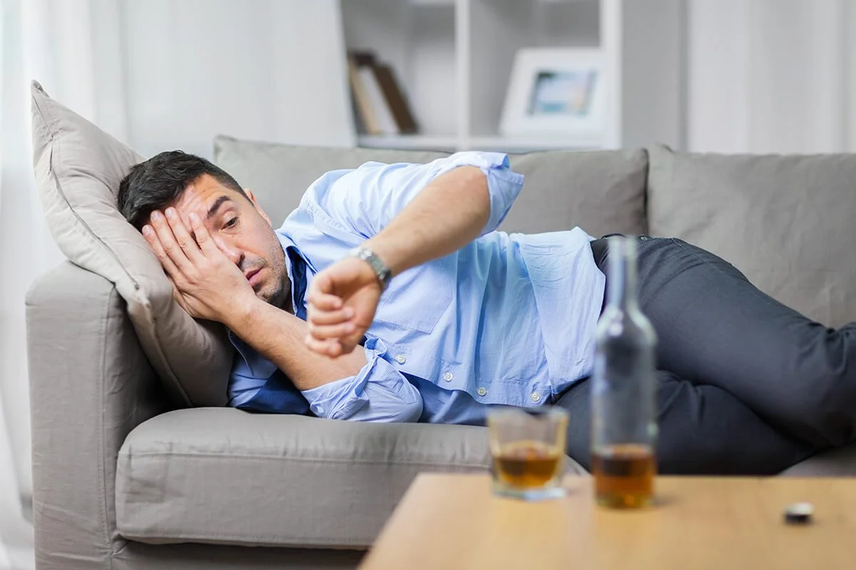 A man lying on a sofa, looking at his watch, with an expression of concern or fatigue, next to beer bottles and glasses on a table.