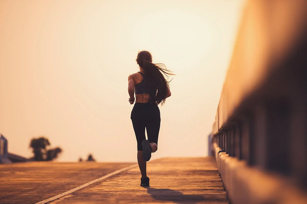 A woman jogging outdoors during sunset or sunrise on an elevated pathway or bridge with a railing on the right side.
