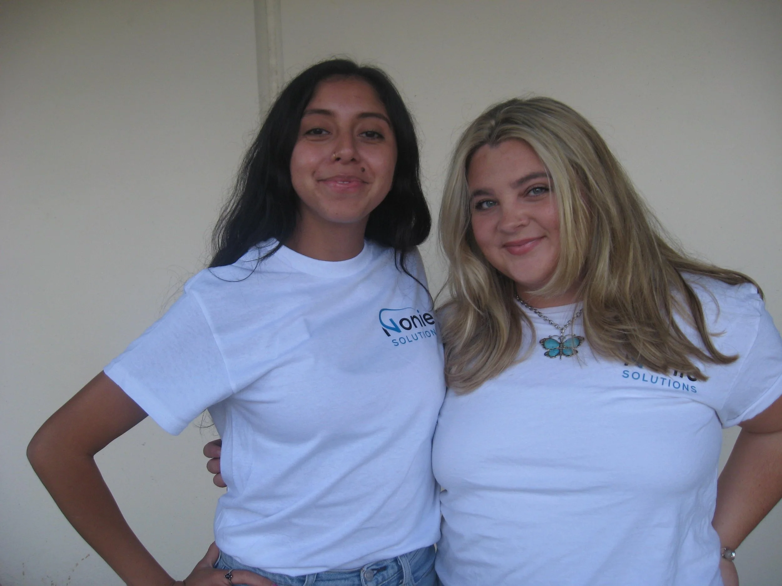 Two women standing side by side, smiling at the camera, wearing white t-shirts with a logo that reads 'Nonie Solutions'.