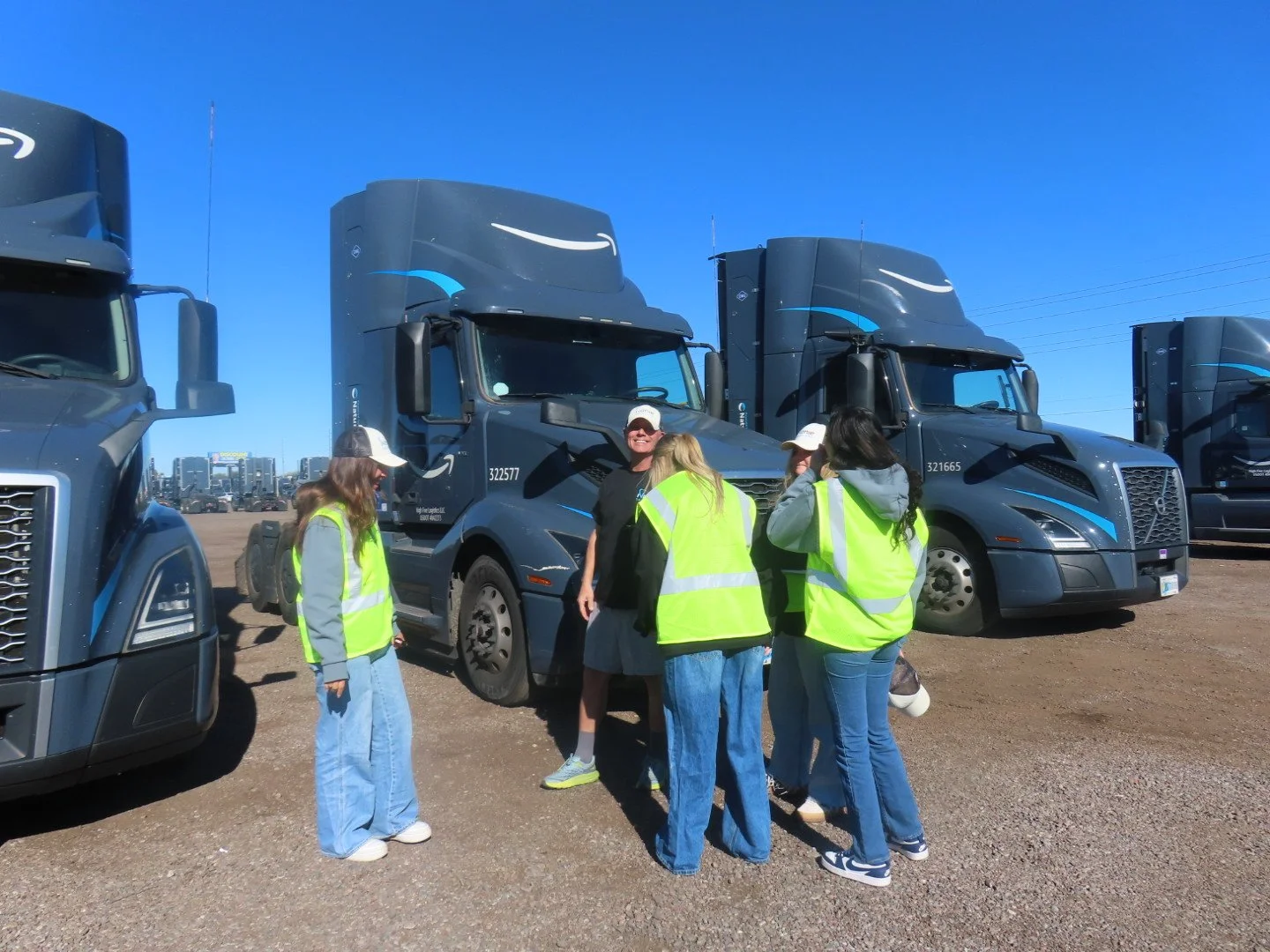 Group of people in yellow safety vests and hats gathered around semi-trucks on a sunny day.