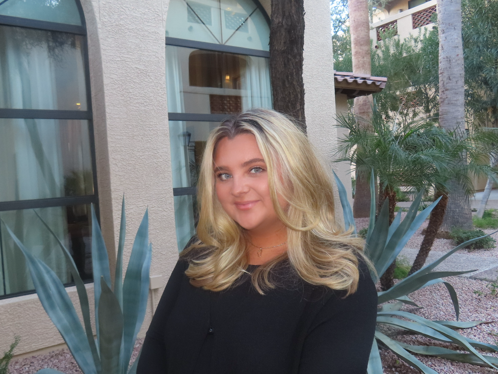 A young woman with blonde hair, wearing a black top, smiling outdoors in front of a building with large windows and tropical plants.