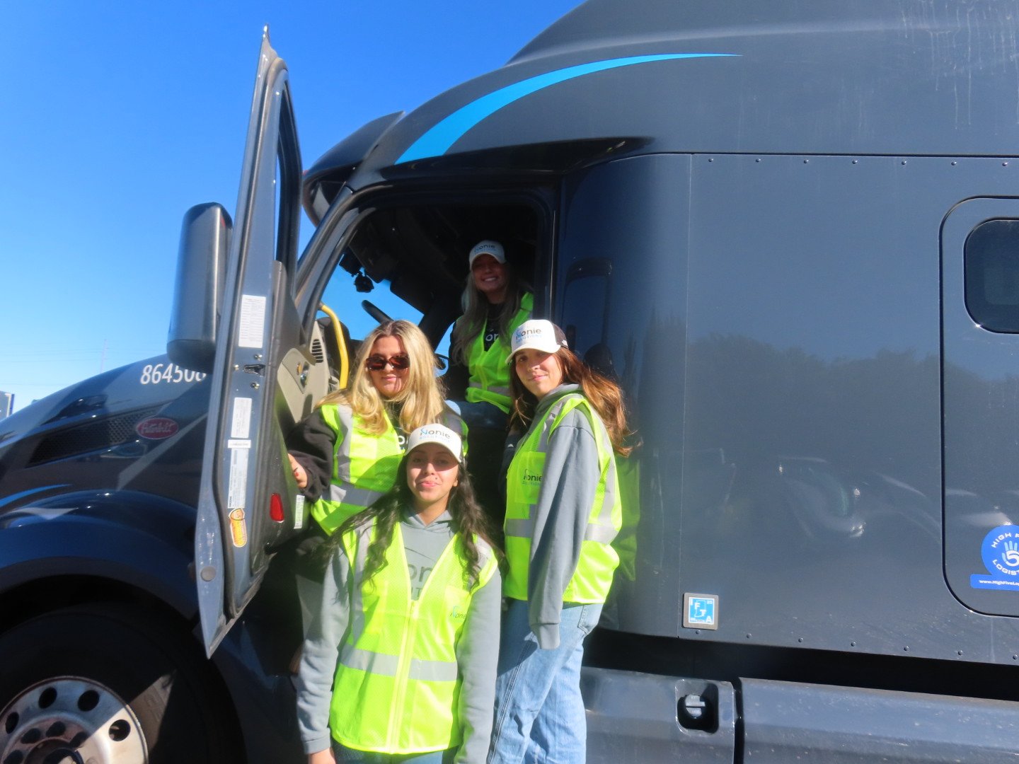 Four women in high-visibility vests and hats standing at the entrance of a large gray semi-truck on a clear day.