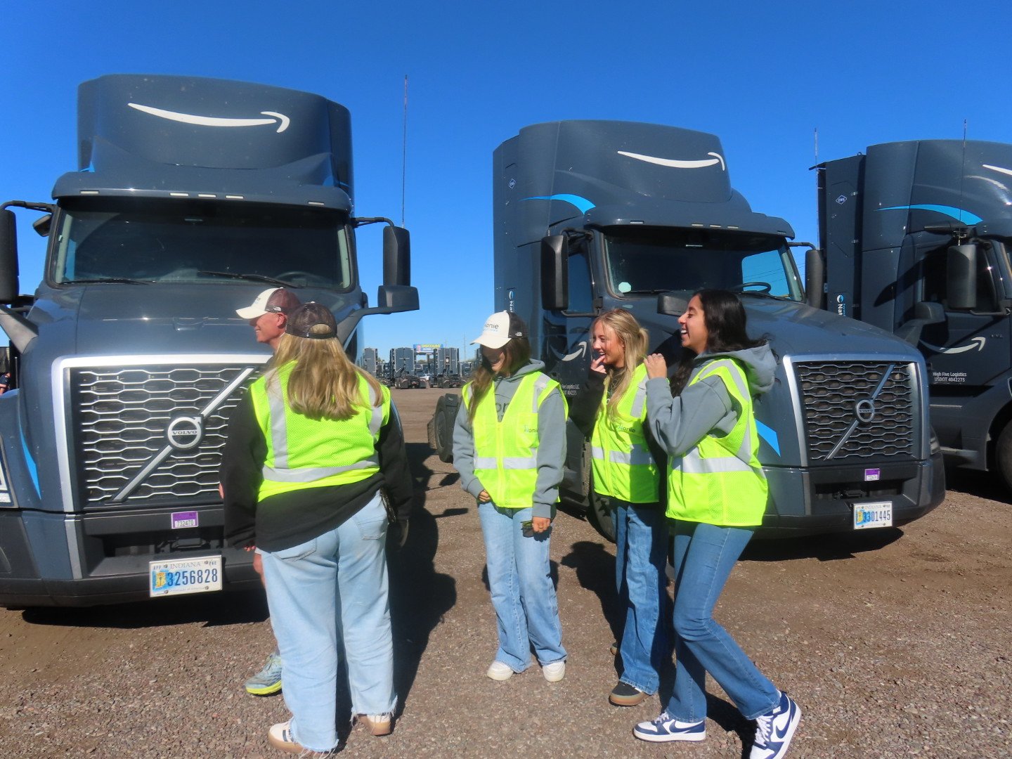 Four women in high-visibility vests standing and laughing in front of two large gray trucks, with a city skyline in the background on a clear day.