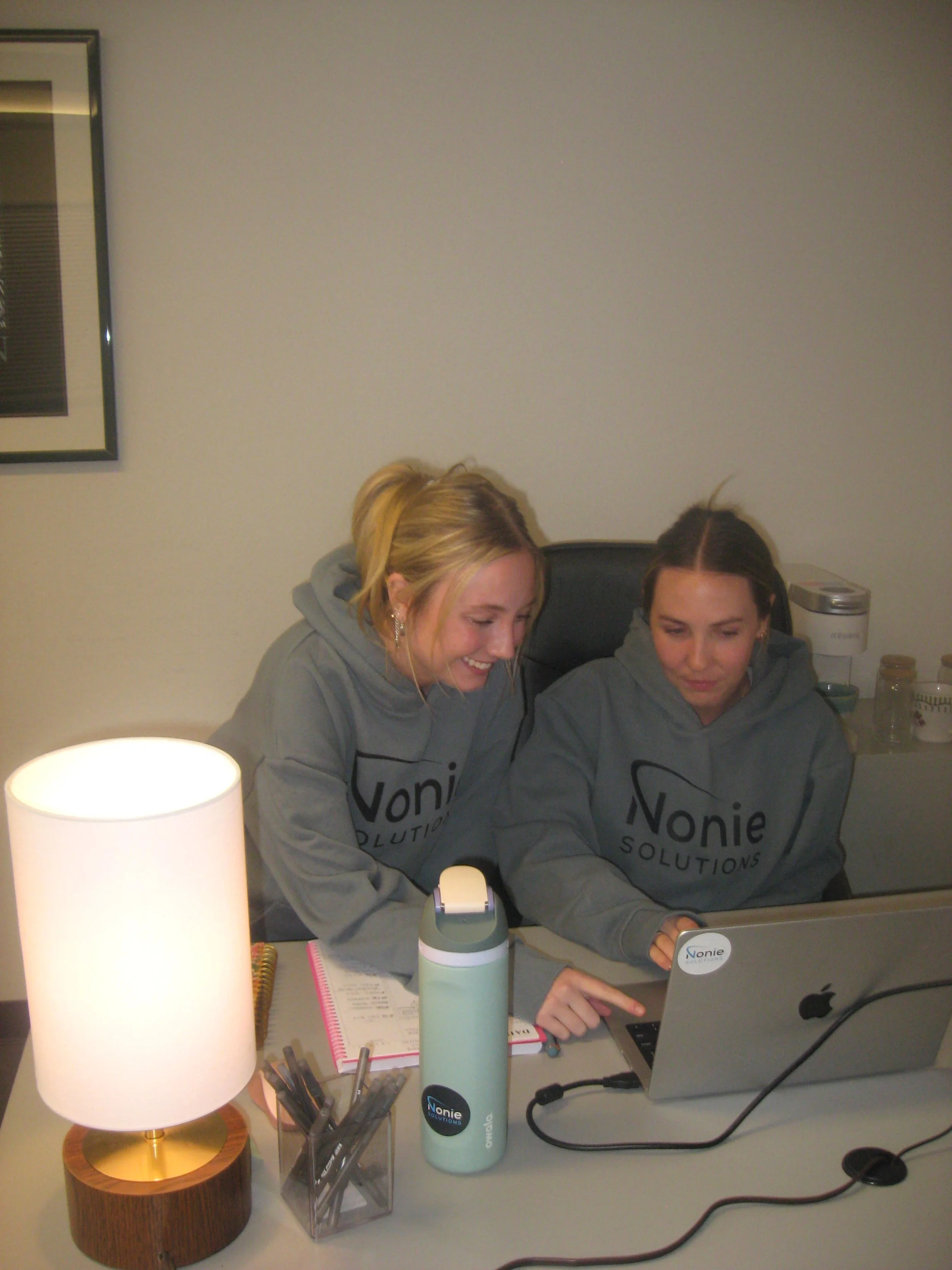 Two women wearing gray hoodies with Nonie Solutions logo, sitting at a desk, looking at a laptop, smiling and pointing at the screen.
