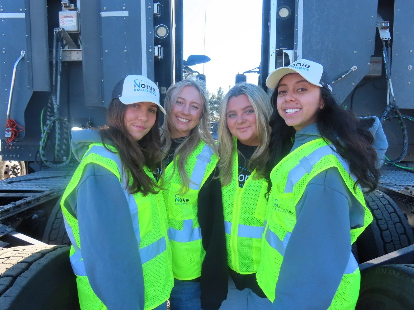 Four young women wearing bright yellow safety vests and white caps with 'Nonie Solutions' logo, standing outdoors in front of large industrial equipment, smiling for the camera.