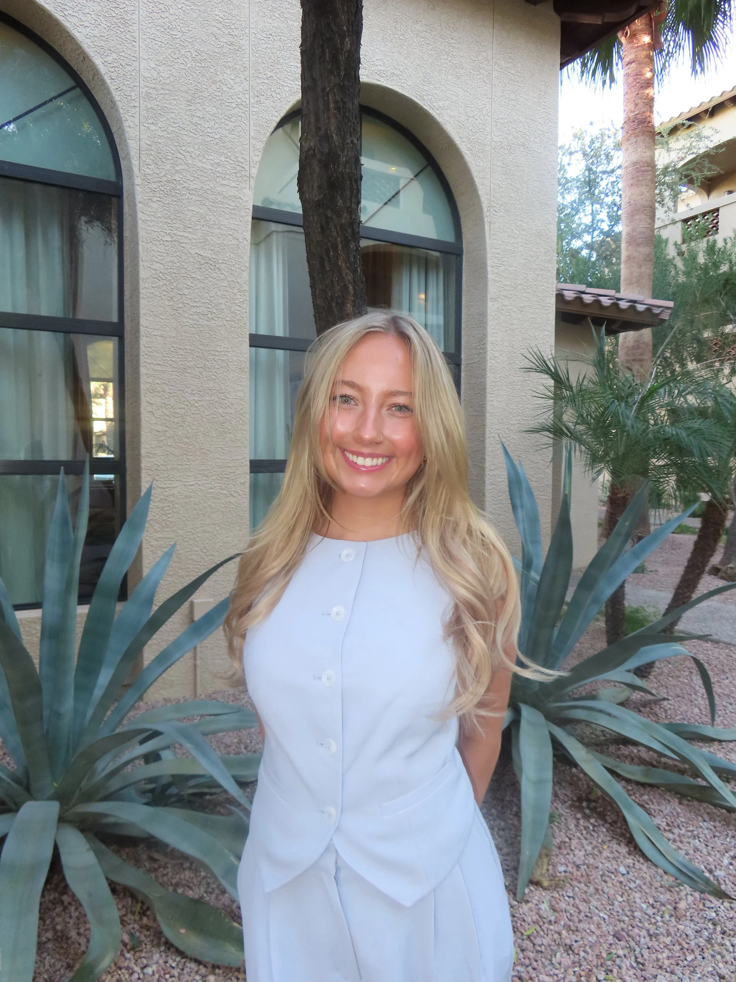 A smiling woman with long blonde hair, wearing a light-colored sleeveless dress, standing outdoors in front of desert plants and a beige stucco building with arched windows.