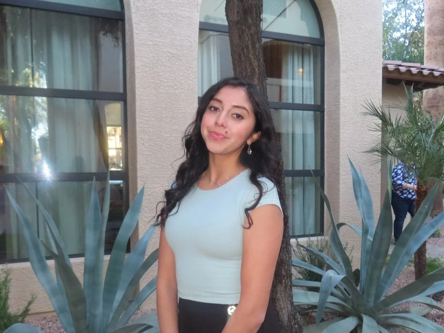 A young woman with long dark curly hair wearing a white top and earrings standing outdoors near a tree, with plants and a building in the background.