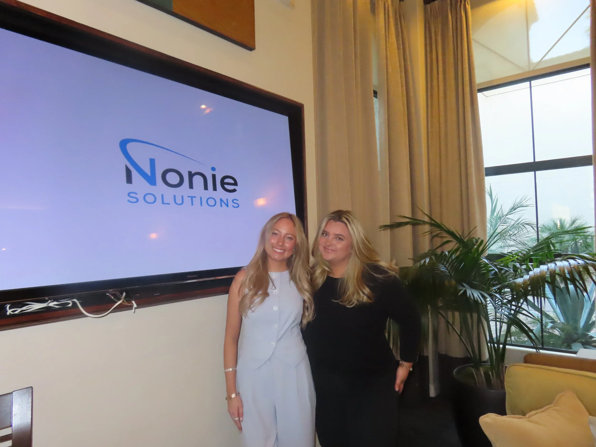 Two women smiling and standing together in a room with curtains and a large window; one woman is wearing a white outfit, and the other is wearing black; there is a large plant and a yellow sofa in the background; a large screen displays the logo "Nonie Solutions."