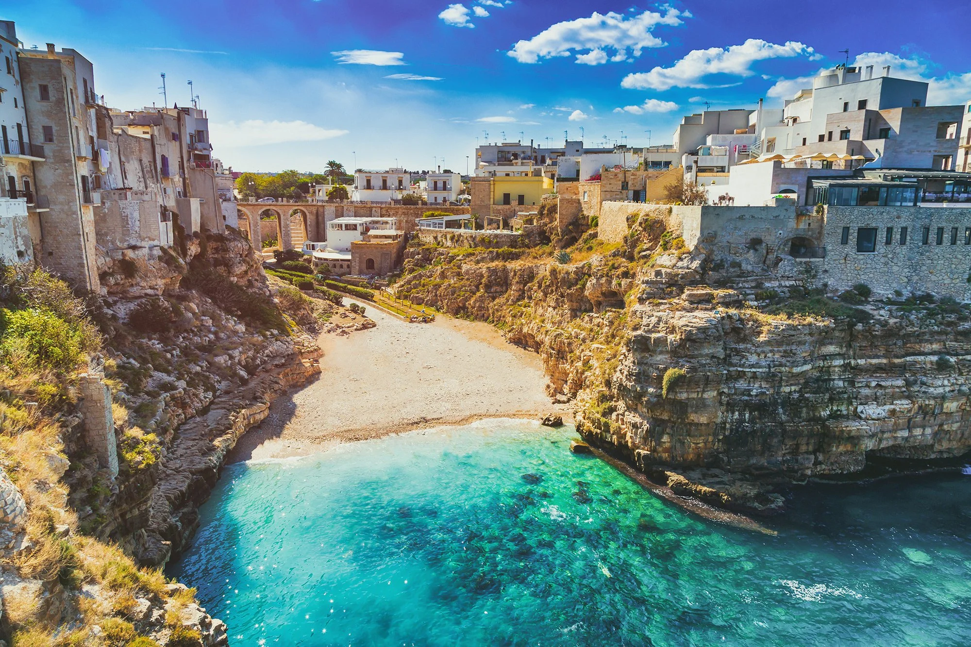 Coucher de soleil sur une côte rocheuse avec des bâtiments blancs en terrasses, une plage de galets et une mer turquoise calme.