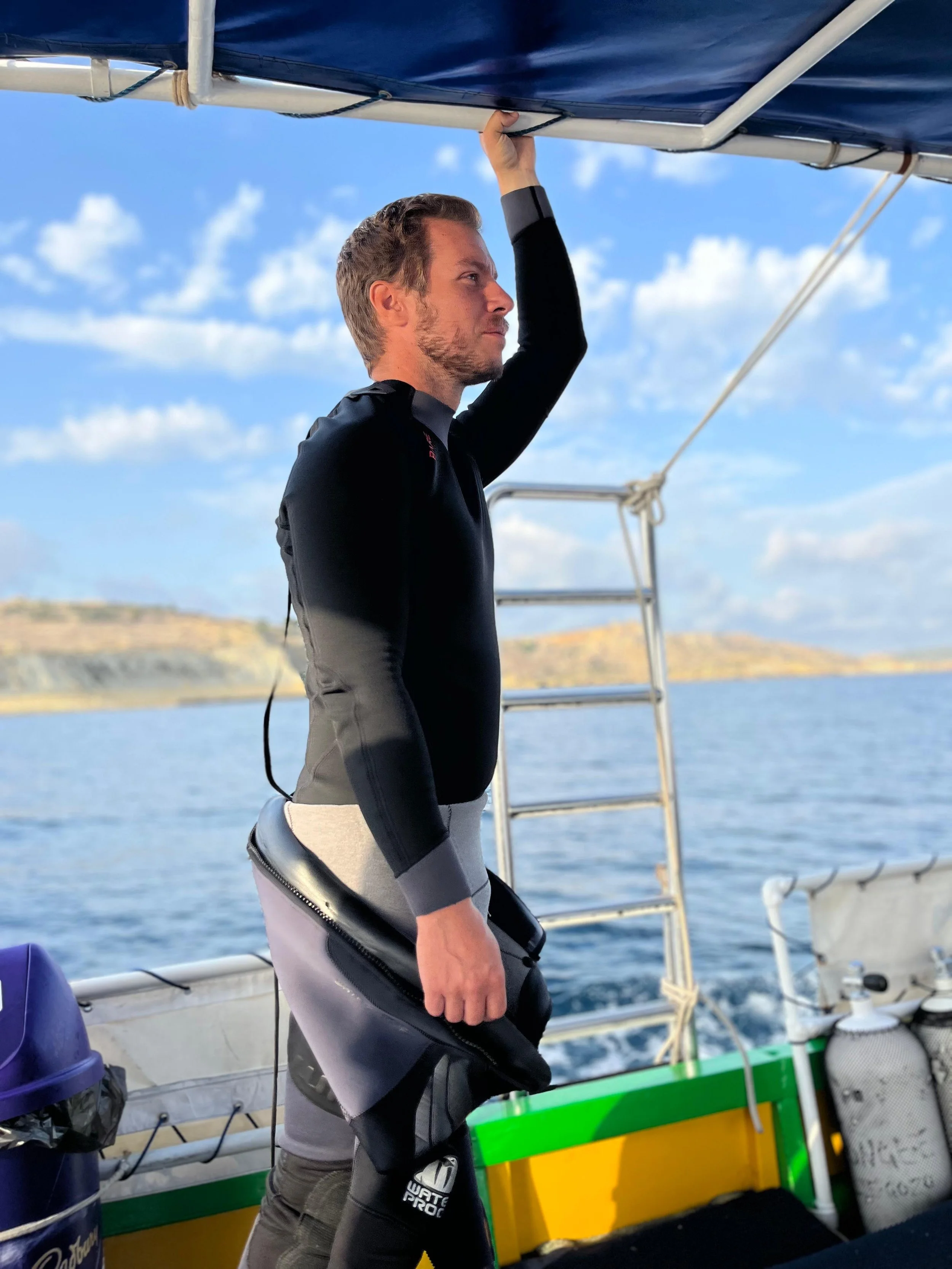 Un homme en vêtements de plongée sur un bateau regarde au loin, tenant une barre ou un support au-dessus de sa tête, avec la mer et un ciel bleu avec quelques nuages en arrière-plan.