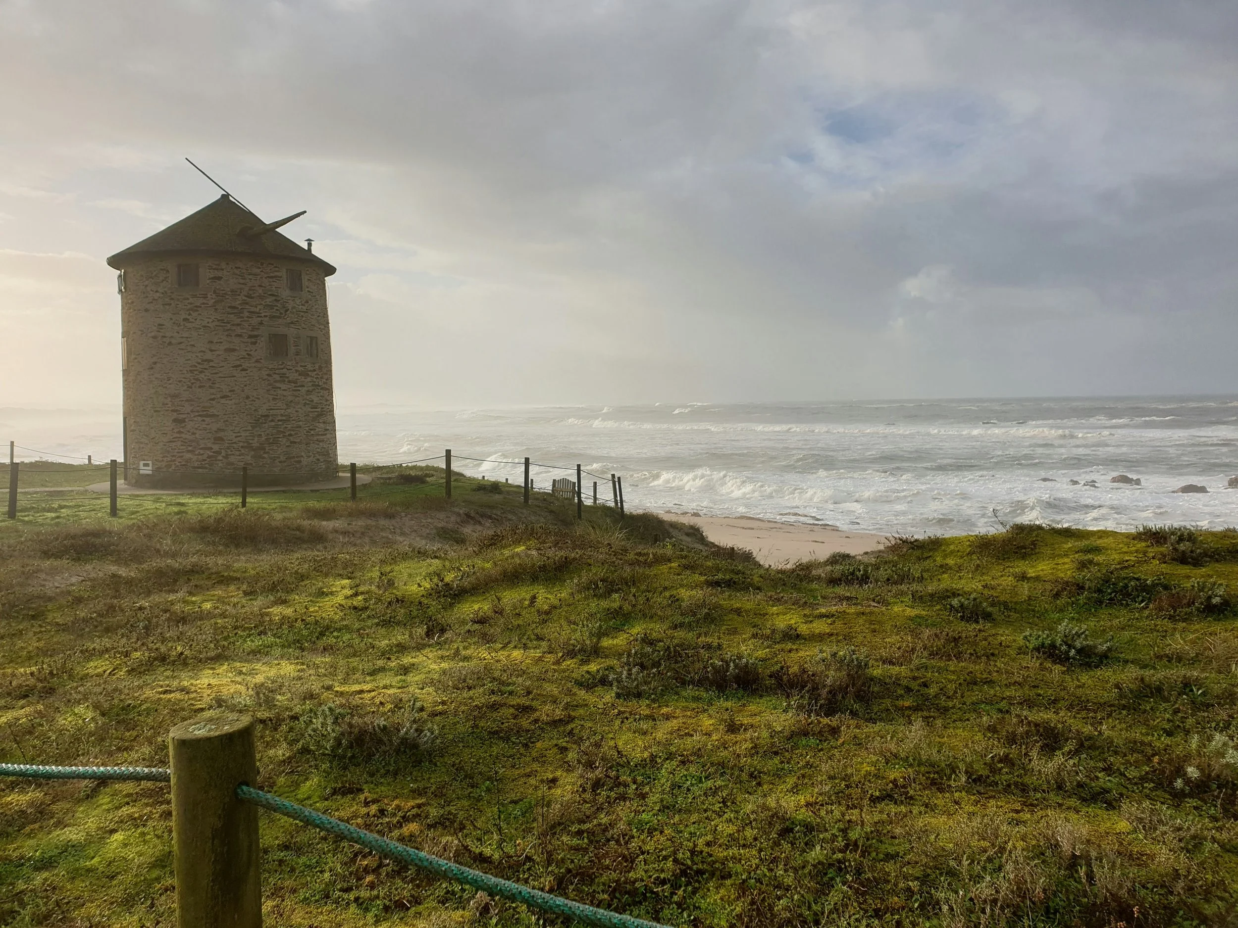 Tour bretonne en pierre sur la côte avec la mer agitée en arrière-plan, ciel nuageux et végétation verte au premier plan.