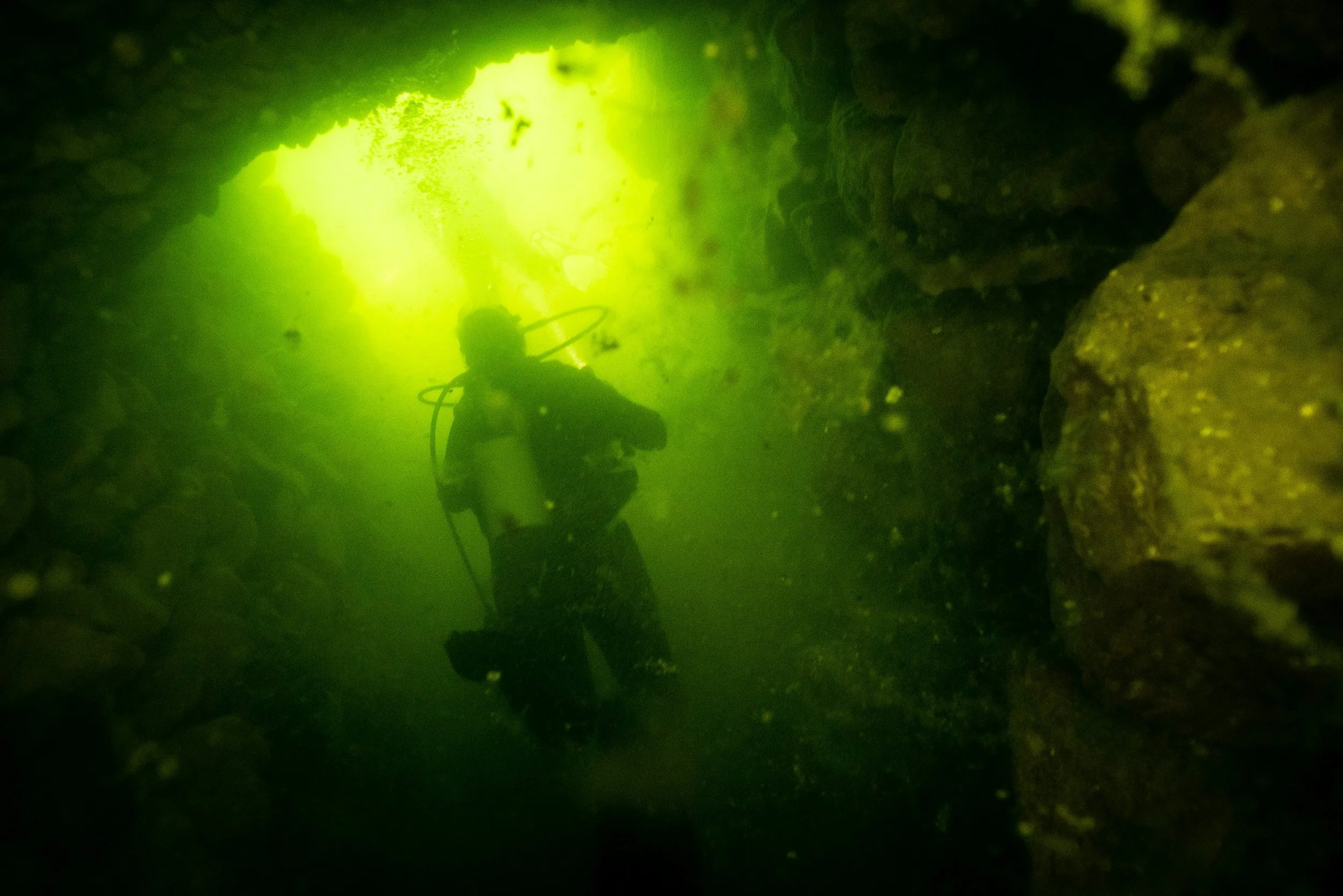 Scuba diver exploring underwater cave with greenish water and rocks.