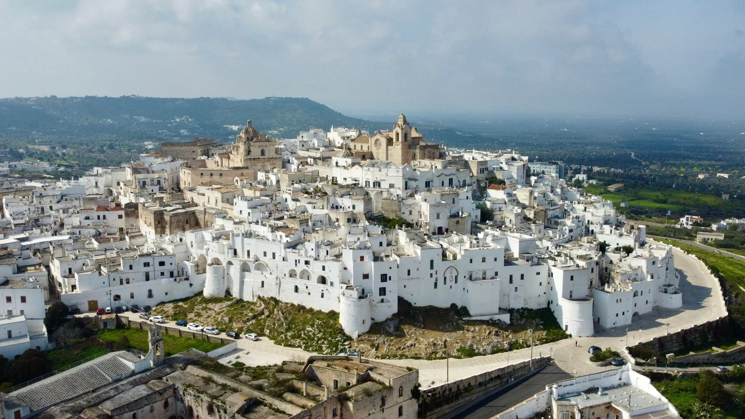 Ville blanche perchée en hauteur avec des bâtiments en pierre, une église au centre dominateur, entourée de remparts, sous un ciel nuageux, avec un paysage rural en arrière-plan. Italie du sud, les pouilles