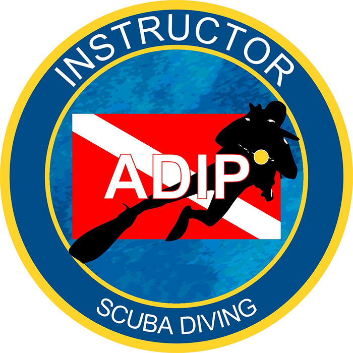 Logo circulaire avec un bord bleu et une bordure jaune. En haut, écrit "INSTRUCTOR" et en bas, "SCUBA DIVING". Au centre, fond bleu avec un drapeau diagonal rouge et blanc. Sur ce fond, silhouette d'un requin noir avec un plongeur noir portant un masque et un tuba. La lettre "ADIP" en blanc est superposée sur le requin et le plongeur.