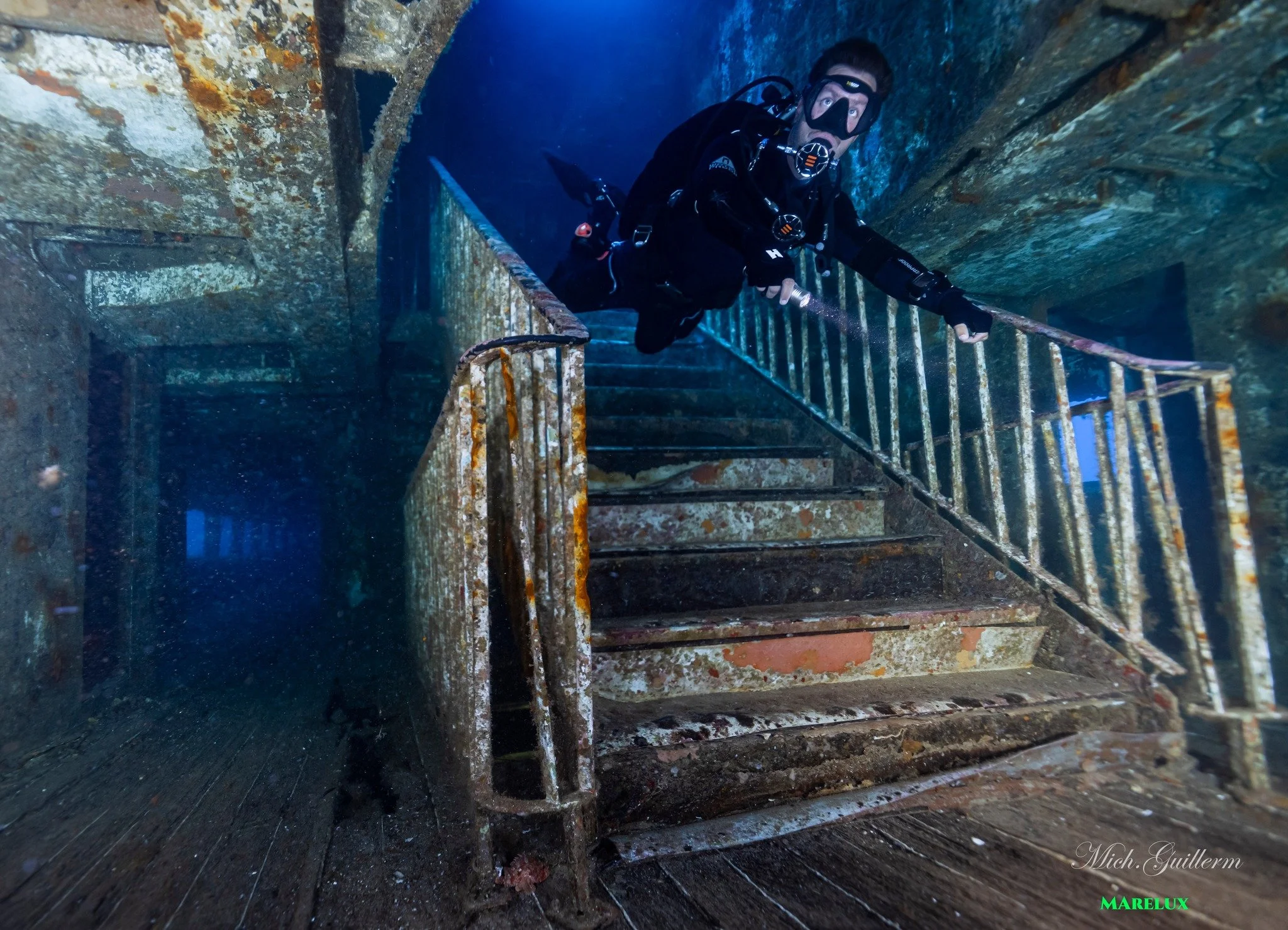 Plongeur explorant un site sous-marin avec une structure en métal rouillée, probablement un navire ou un bâtiment submergé, dans une ambiance sombre et mystérieuse.