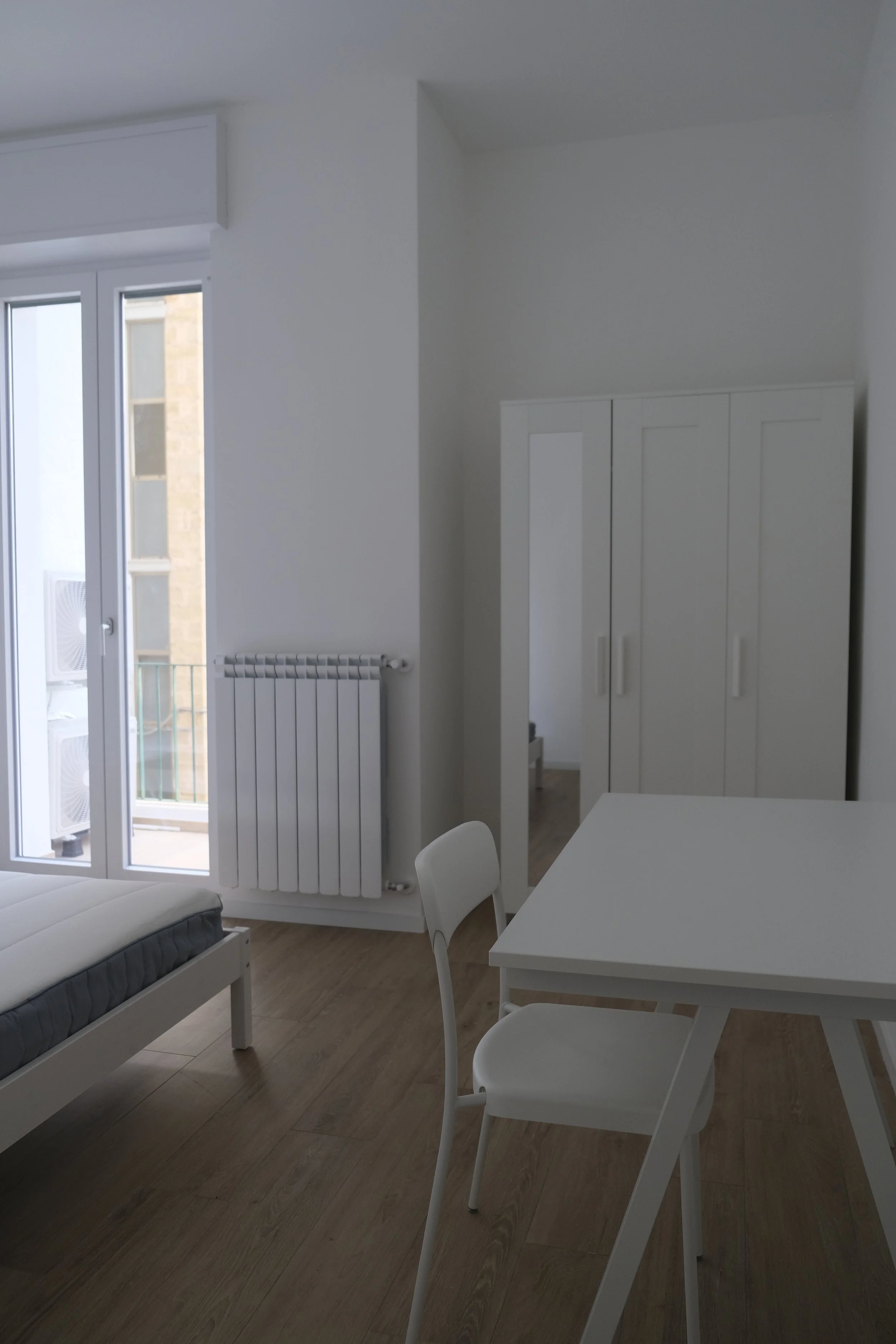 Intérieur d'une chambre blanche, avec un lit simple, une table blanche, une chaise, un placard, une radiateur et une porte-fenêtre menant à un balcon avec des unités de climatisation extérieures.