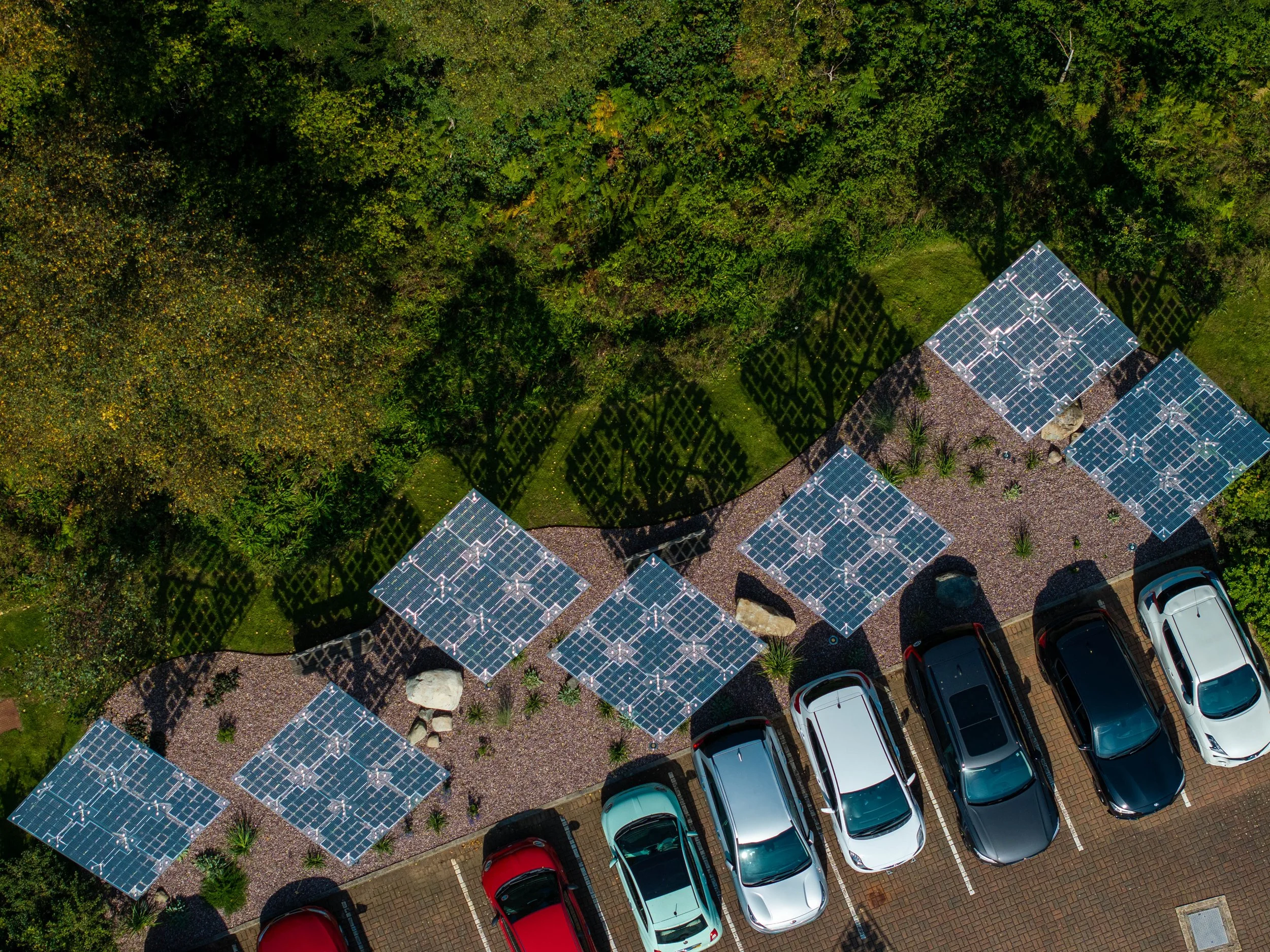 Parking lot with several cars and solar panel arrays on a landscaped area surrounded by trees.