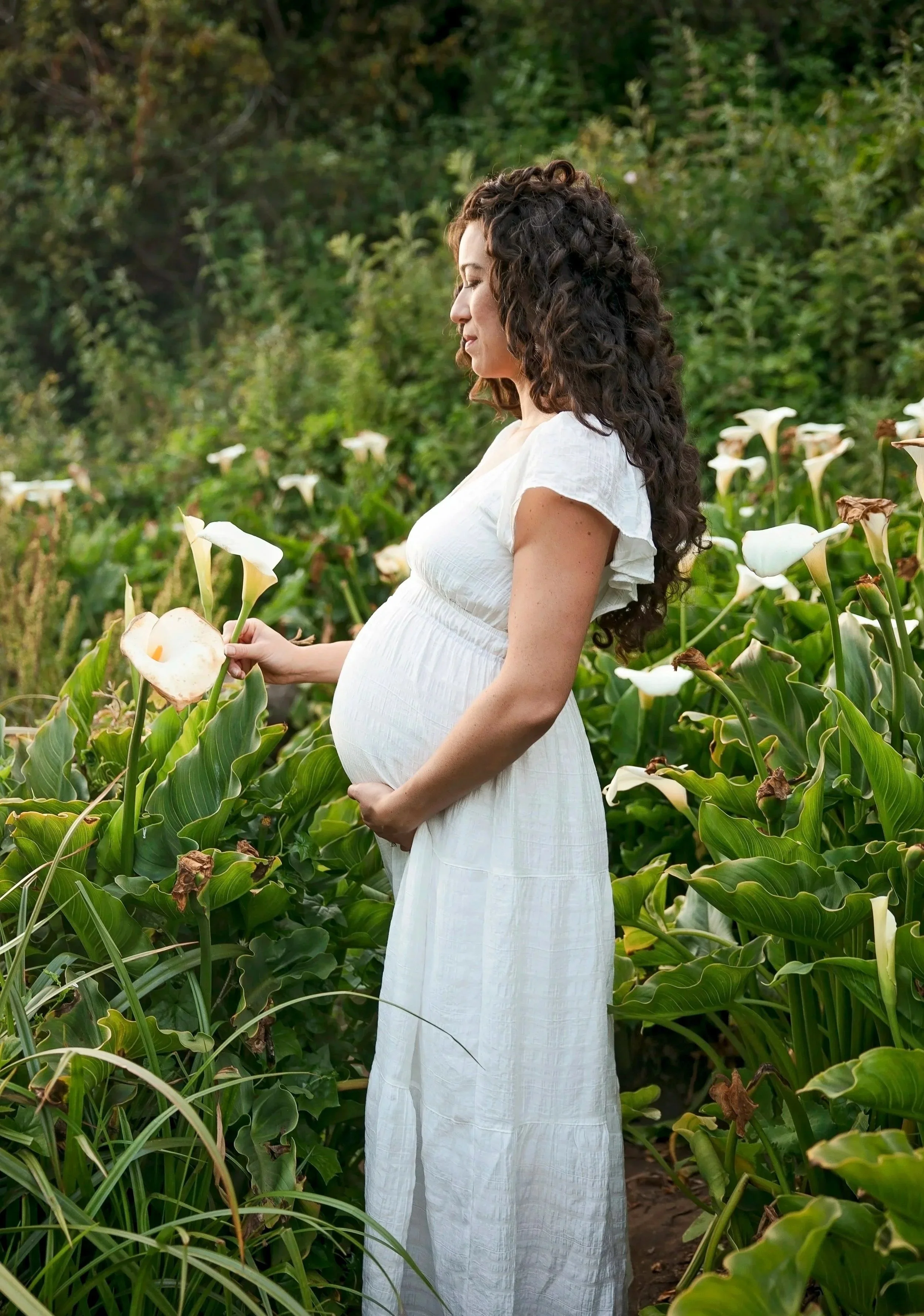 Pregnant woman in white dress standing among lush green plants and white calla lilies, holding a calla lily flower, with a thoughtful expression and eyes closed.