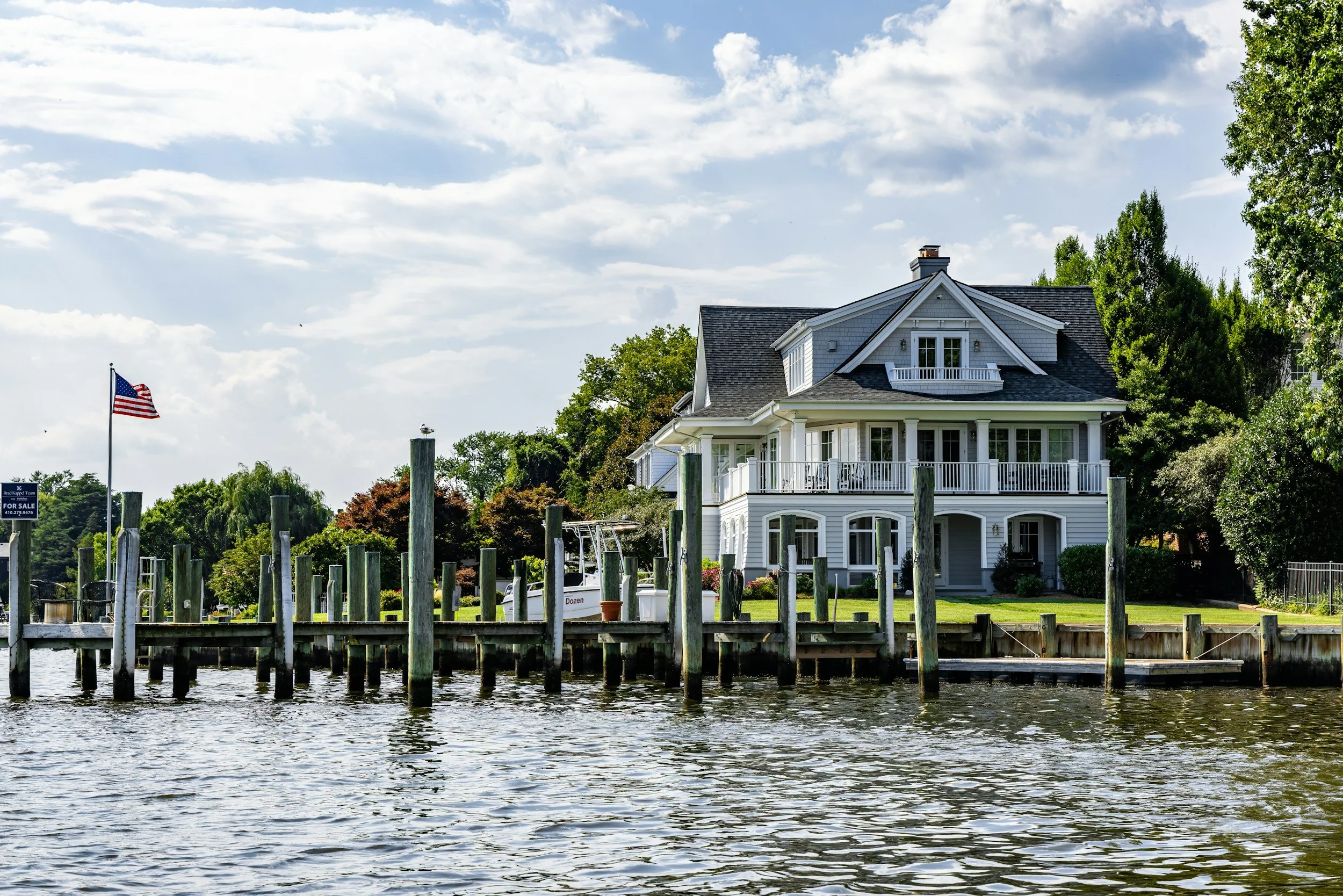 A white multi-story house with a large porch, located by a river, surrounded by green trees, with an American flag on a flagpole, under a partly cloudy sky.