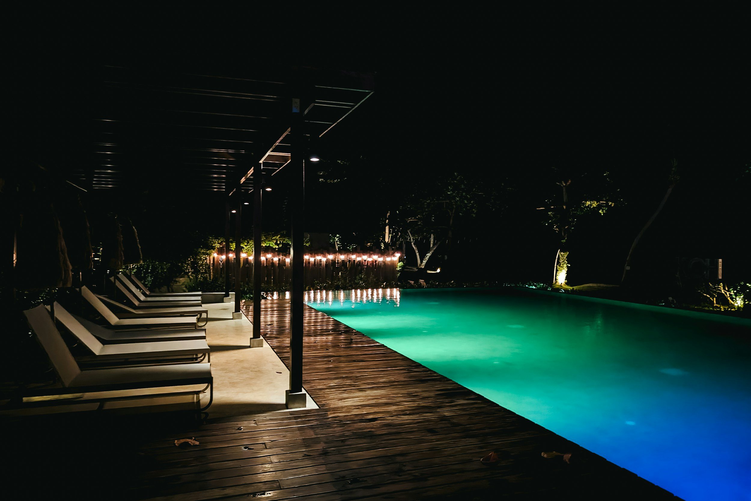 Night view of an illuminated swimming pool with lounge chairs along the deck, surrounded by trees and soft outdoor lighting.