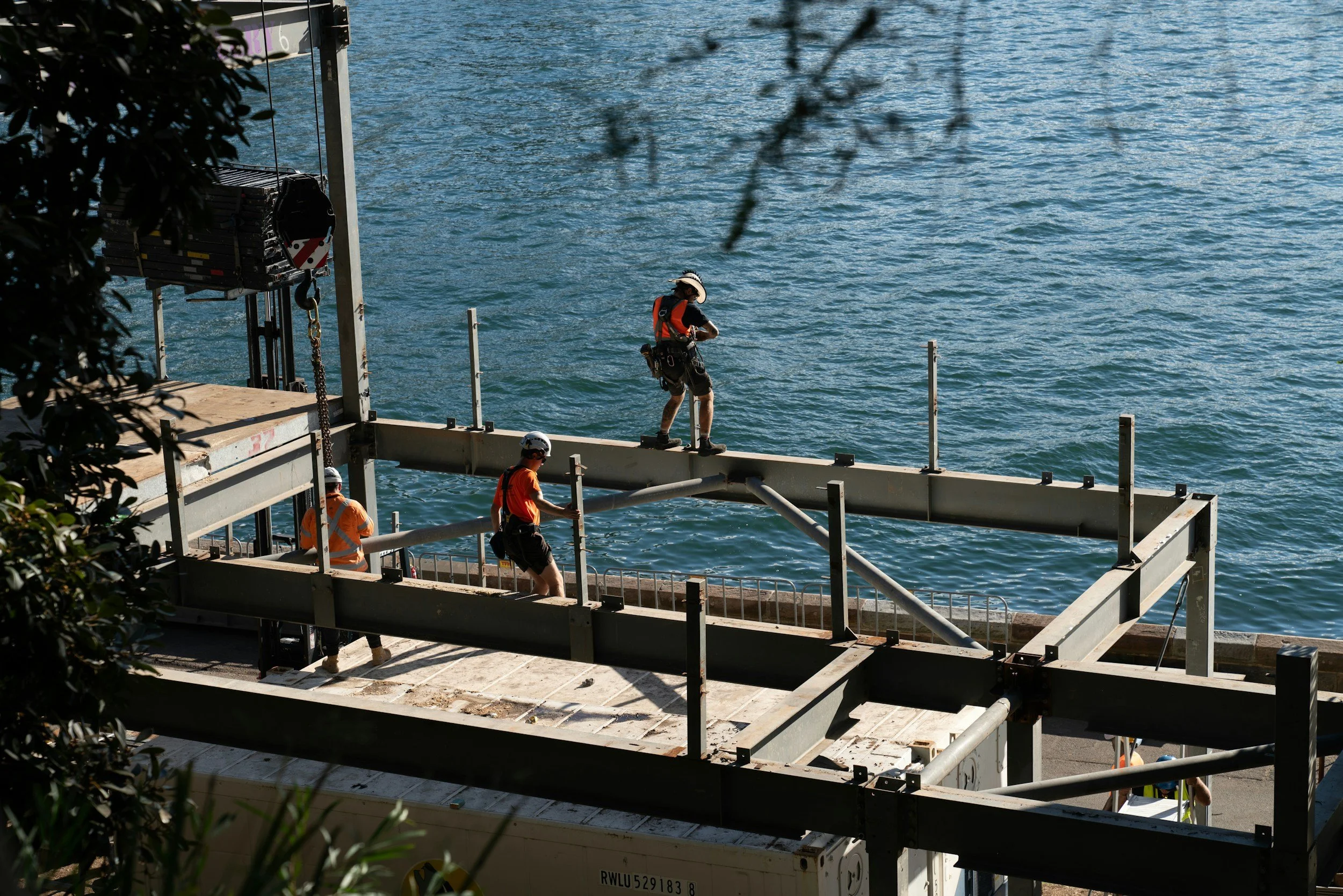 Workers in safety gear working on a construction site near a body of water, with steel framing and equipment.