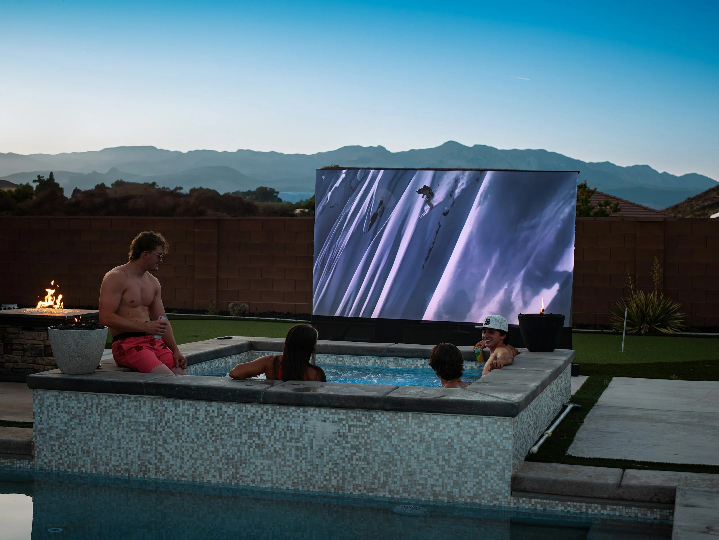 People relaxing in a backyard hot tub watching a movie on an outdoor screen with mountain scenery in the background during dusk.